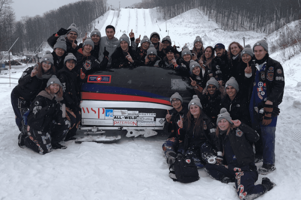 large group of students posing with toboggan outdoors in snowy area